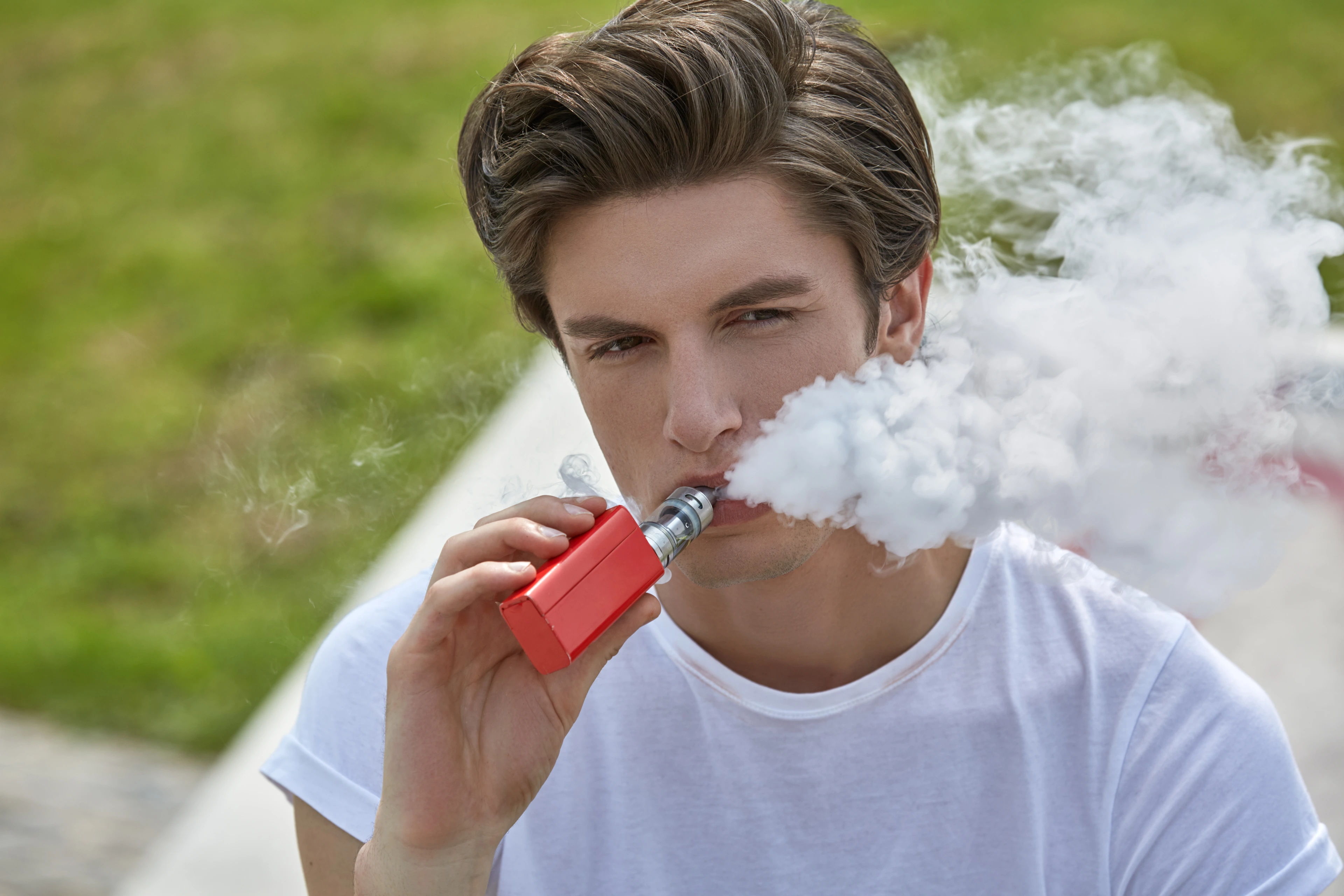 Junger Mann in T-Shirt und Jeans sitzt e-Zigarette rauchend im Sonnenschein und bläst lässig eine weiße Dampfwolke.