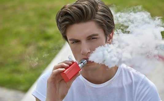 Junger Mann in T-Shirt und Jeans sitzt e-Zigarette rauchend im Sonnenschein und bläst lässig eine weiße Dampfwolke.