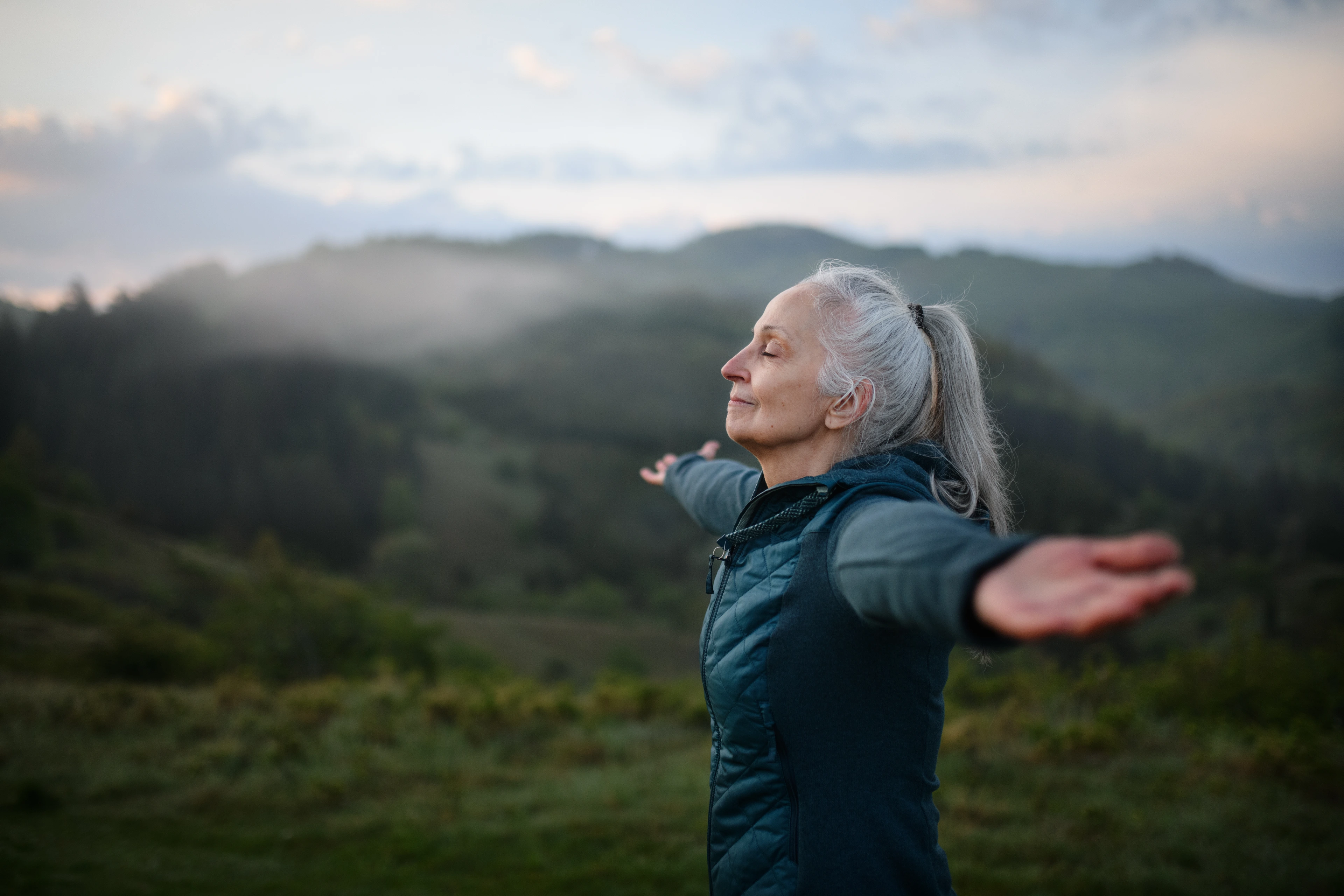 Senior woman doing breathing exercise in nature on early morning with fog and mountains in background.