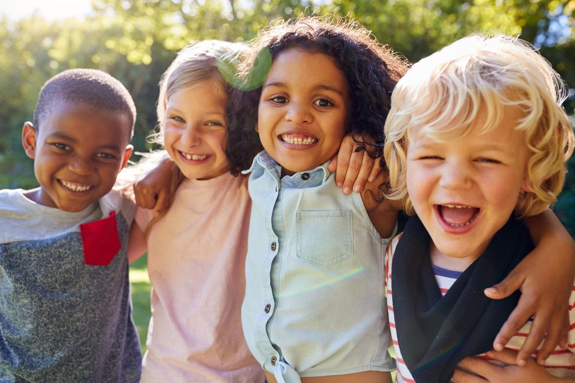 Vier Kinder stehen Arm in Arm im Freien, lachen fröhlich und schauen in die Kamera.
