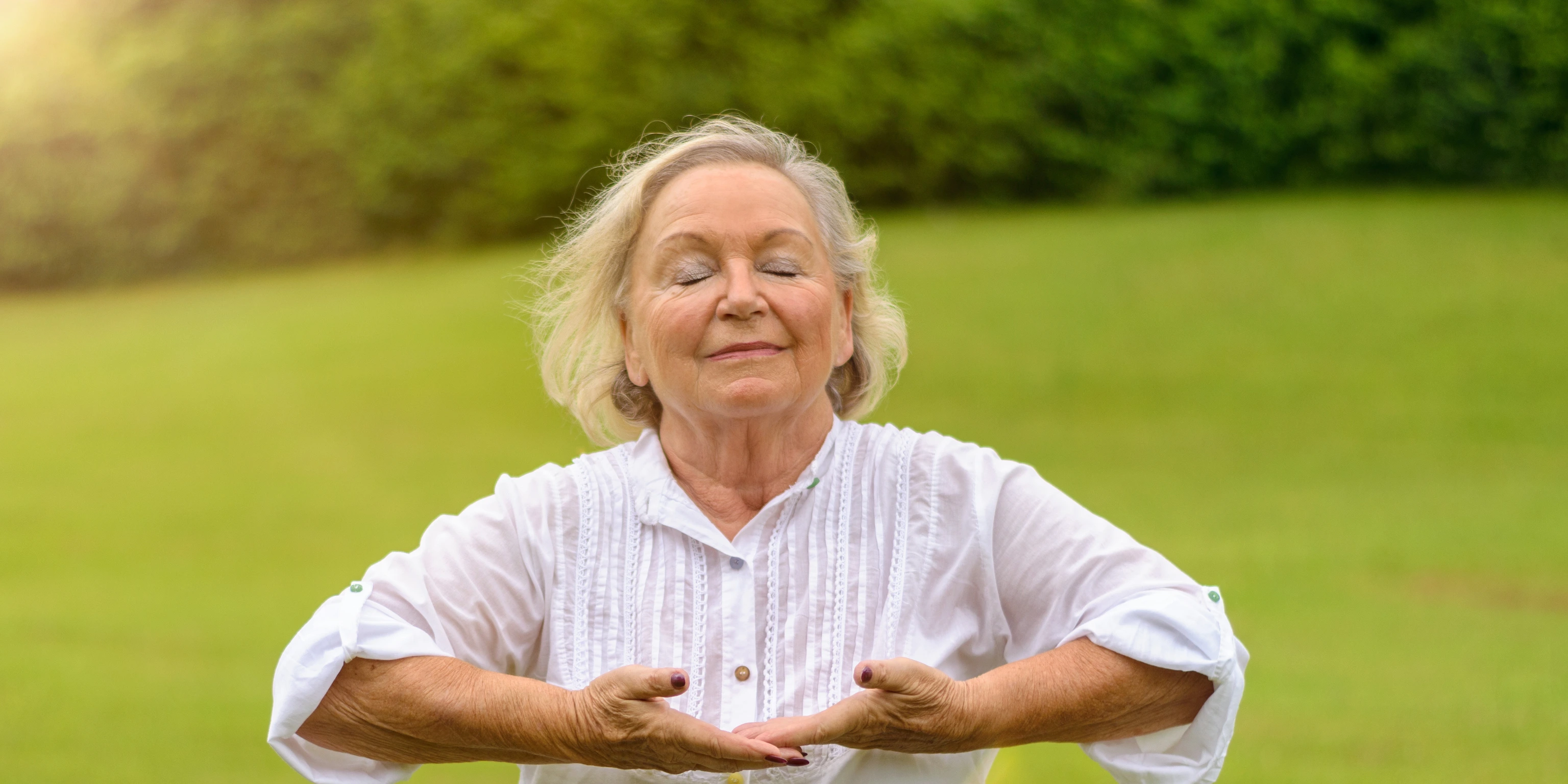 Single beautiful senior woman in white rolled up long sleeve blouse practicing breathing exercises outside with eyes closed on green grass with copy space