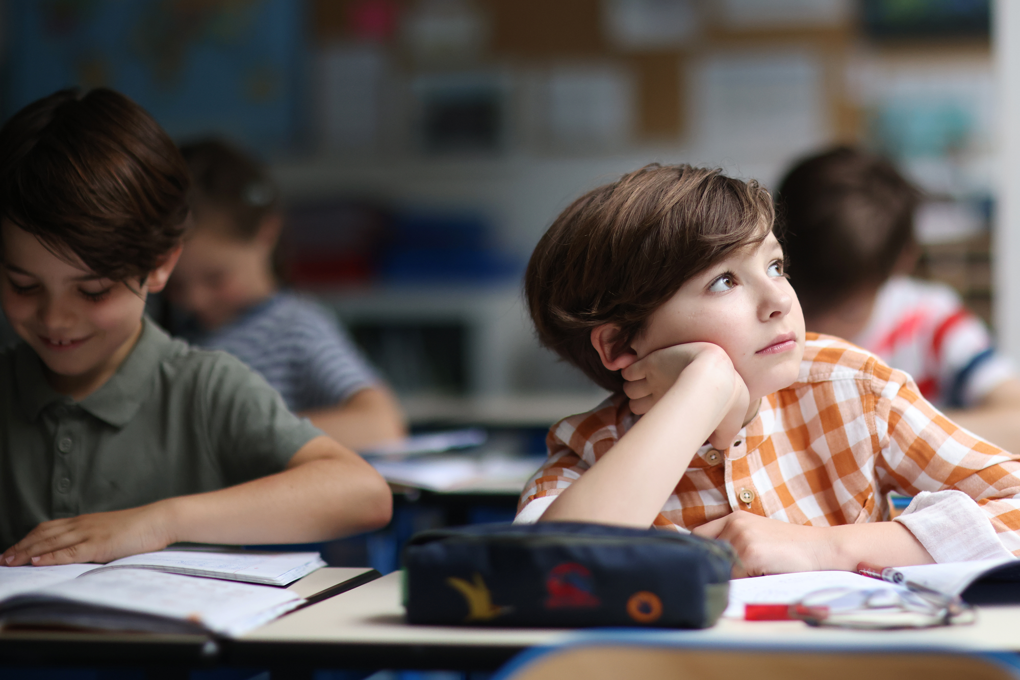 Grundschüler sitzt im Klassenzimmer am Tisch, stützt nachdenklich den Kopf auf die Hand und blickt zur Seite.