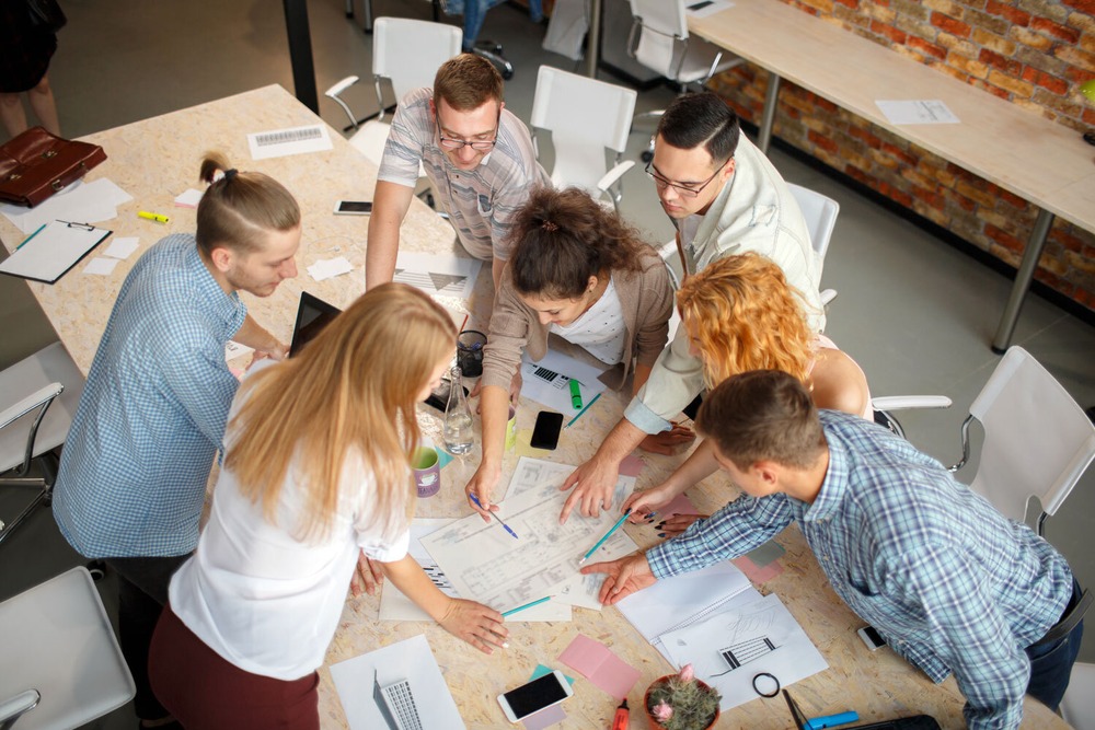 Eine Gruppe junger Menschen über einen Arbeitstisch gebeugt, auf dem verschiedene Arbeitsmaterialien, Papiere liegen