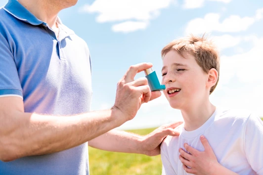 child using inhaler for asthma outside in a park with his father