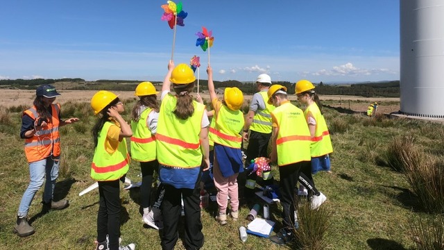 A group of children wearing hard hats and bright safety vests stand together on open grassy land, holding up colourful pinwheels. An adult in an orange safety vest stands nearby giving instructions, and notebooks and equipment are scattered on the ground. The sky is clear and blue above the wide landscape.