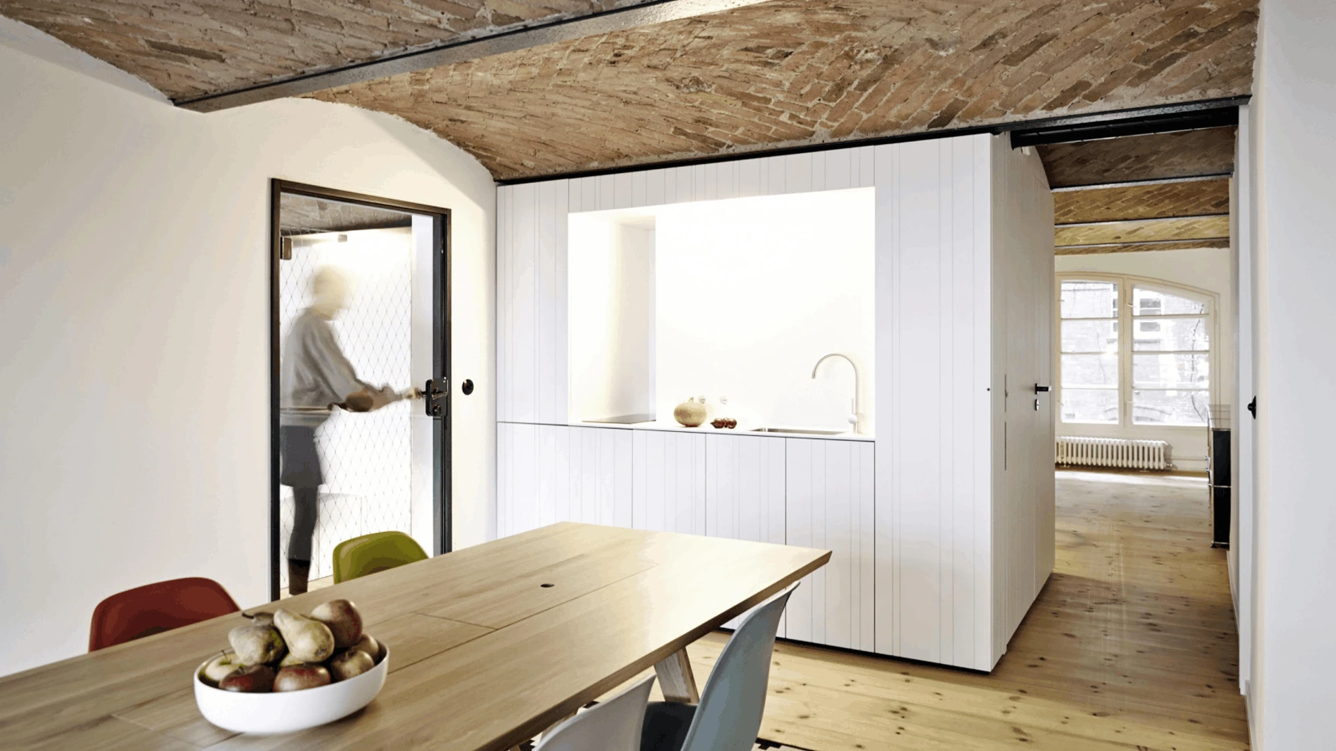 Heller Raum mit Holztisch, bunten Stühlen, PALMBERG-Küchenmodul und einer verschwommenen Person an der Tür.