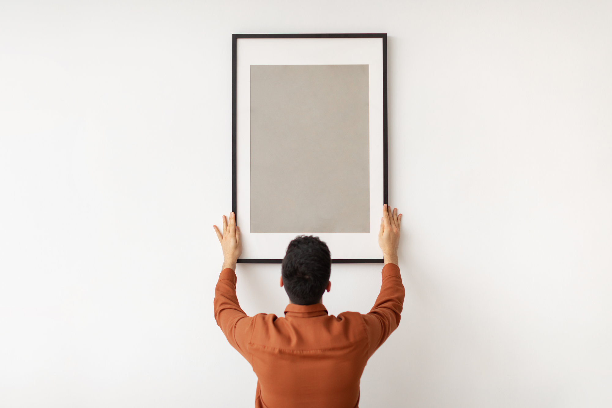 Eine Person hängt ein gerahmtes, schlichtes Bild an einer weißen Wand auf und richtet es mit beiden Händen aus