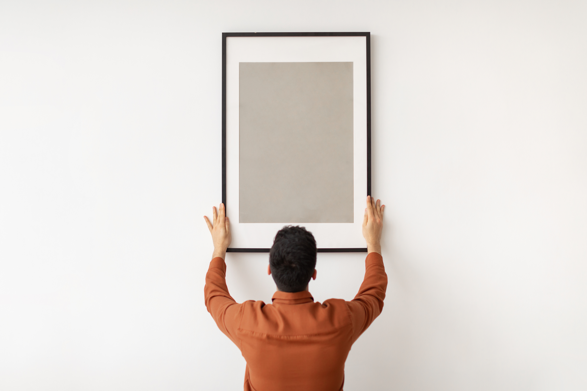 Eine Person hängt ein gerahmtes, schlichtes Bild an einer weißen Wand auf und richtet es mit beiden Händen aus