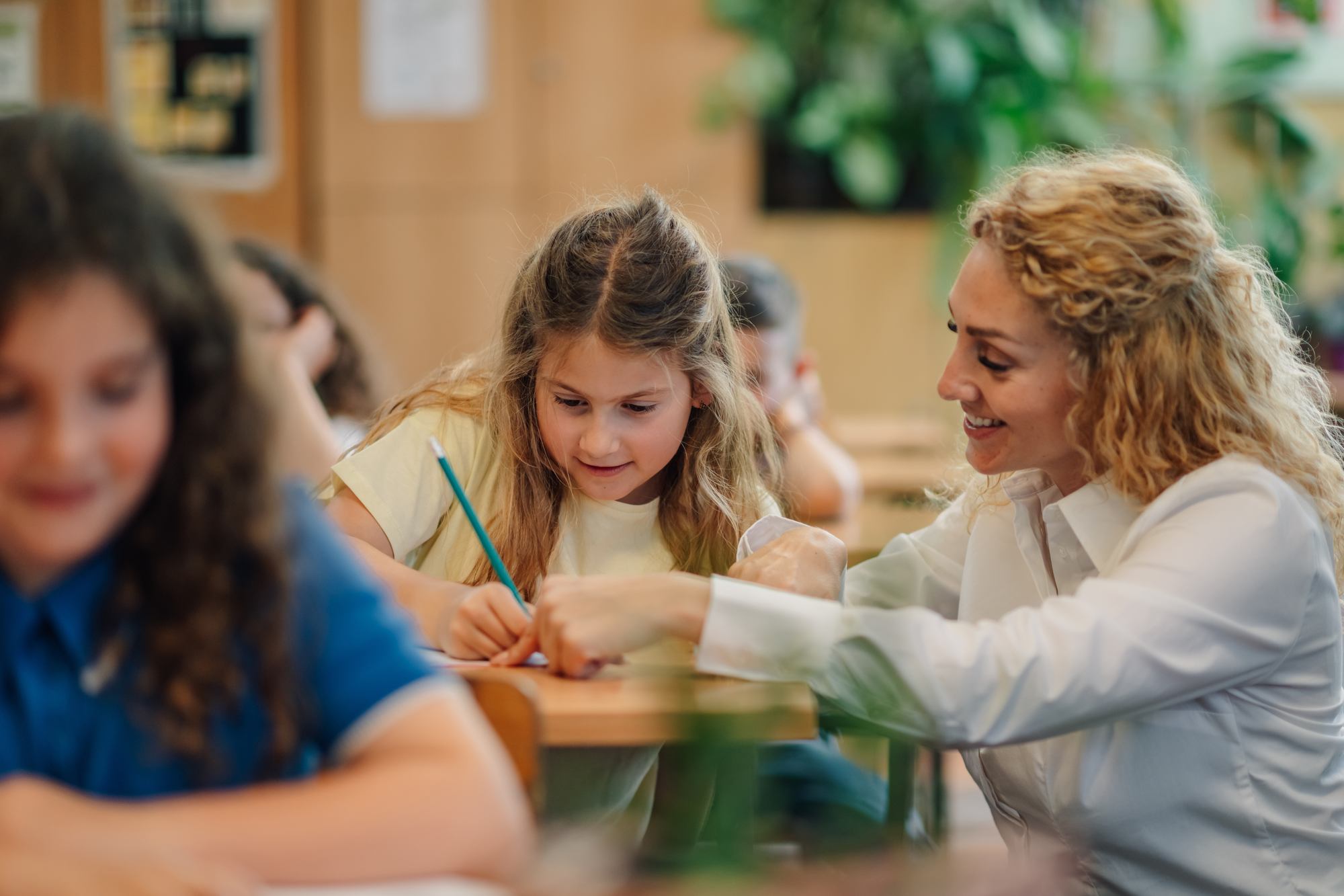 Lehrerin unterstützt ein Schulkind beim Schreiben am Tisch im Klassenzimmer, weitere Kinder arbeiten im Hintergrund.