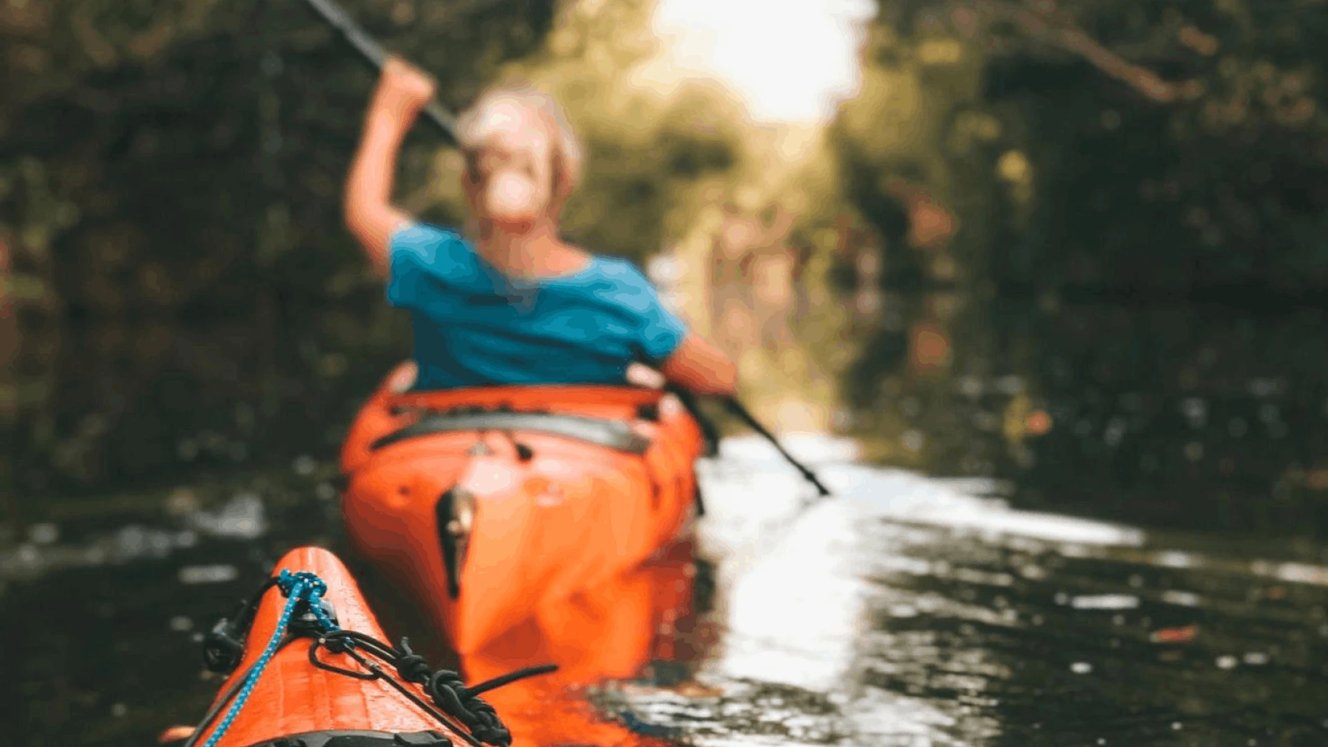 Person paddelt in orange Kajak auf ruhigem Fluss, vordere Bugspitze im Fokus, PALMBERG