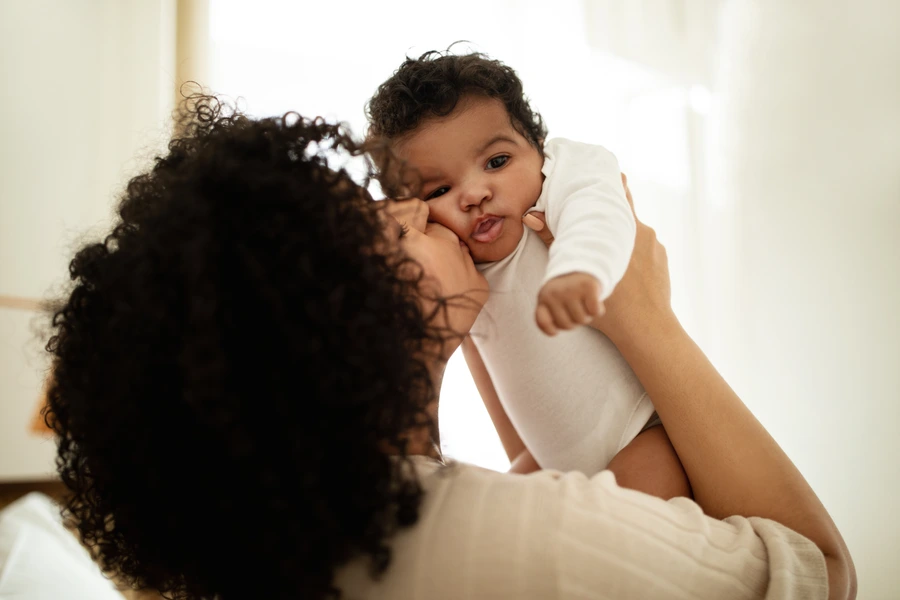 Happy millennial african american mother kissing little child, enjoy relax together, have fun in bedroom interior