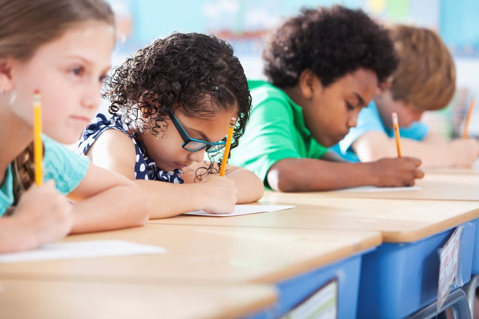 Mehrere Grundschulkinder sitzen nebeneinander an ihren Tischen und schreiben konzentriert mit Bleistiften auf Papier.