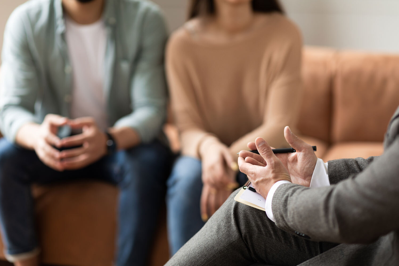 Ein Mann und eine Frau sitzen auf einem Sofa. Im Vordergrund sitzt ein Mann, der die Hände geöffnet auf dem Schoß liegen hat. Es sind keine Gesichter erkennbar.