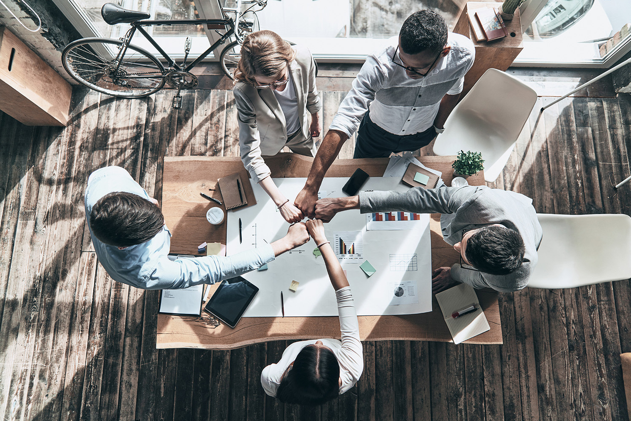 Fünf Geschäftsleute stehen um einen Konferenztisch und stoßen in der Mitte die Fäuste zusammen, Symbol für Teamwork und erfolgreichen Abschluss.