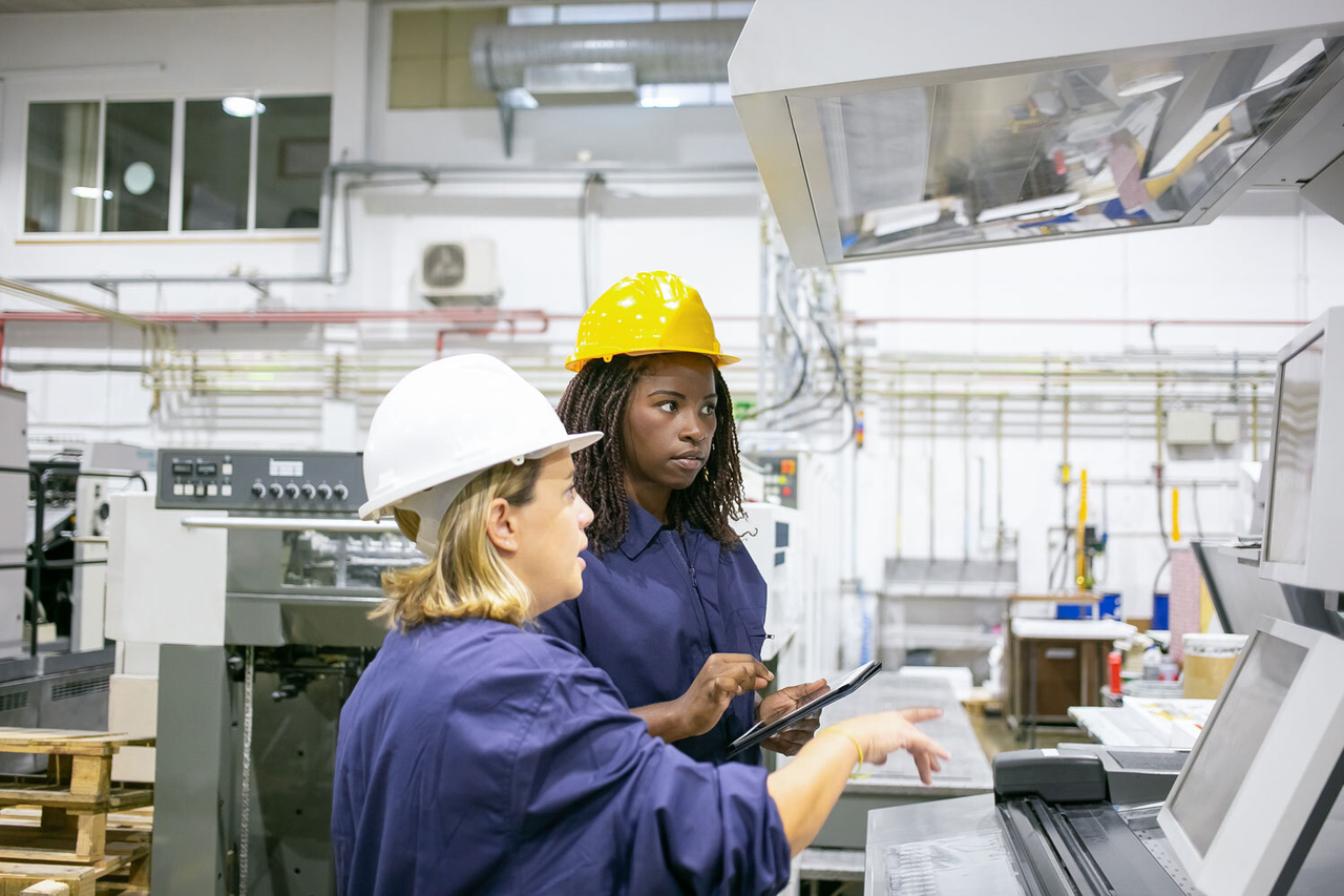 Mentoring auch für Frauen wichtig Ältere erfahrene Arbeitnehmerin erklärt jüngerer Arbeitnehmerin etwas an einer Maschine
