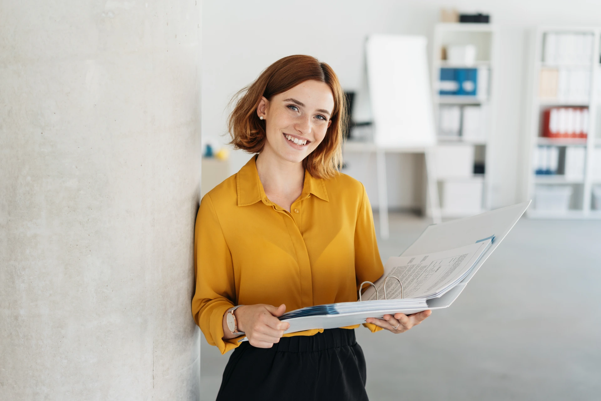 Attractive young office worker holding a large open binder as she looks at the camera with a sweet friendly smile