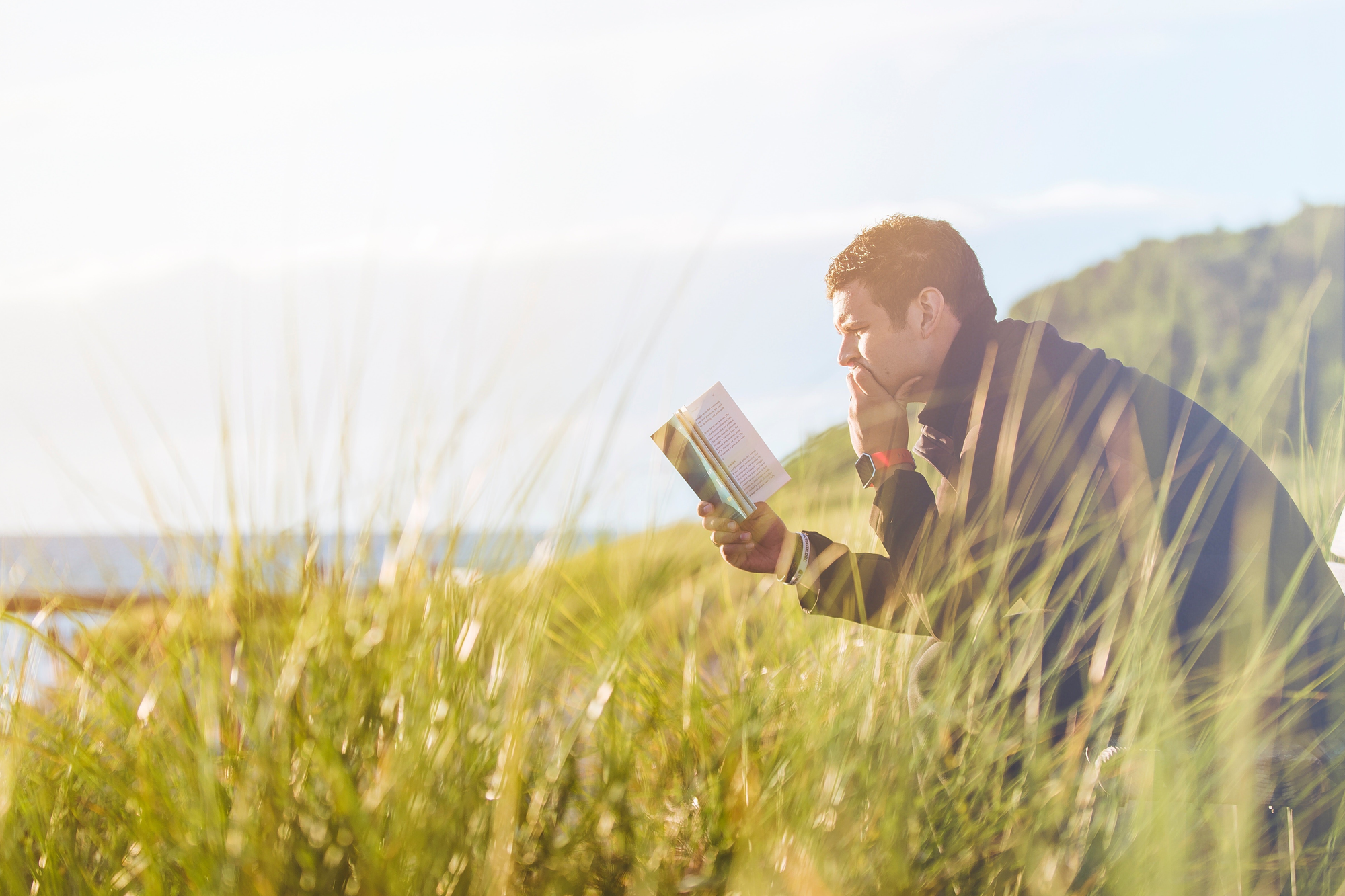 Mann liest ein Buch auf einer Bank am Wasser.