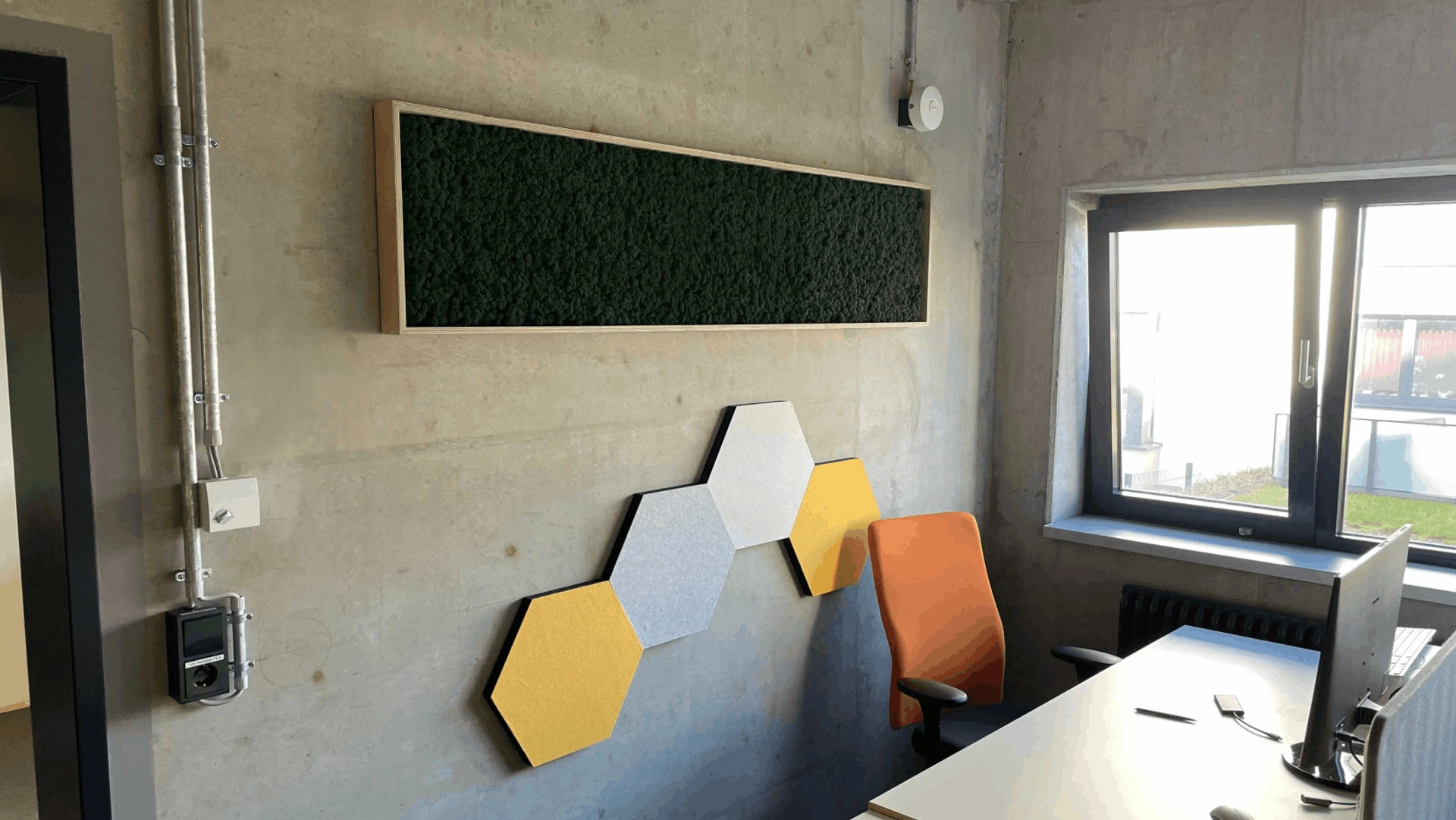 Helles Büro mit PALMBERG-Schreibtisch, orangeem Bürostuhl, sechseckigen Akustikpanelen und grünem Moosbild