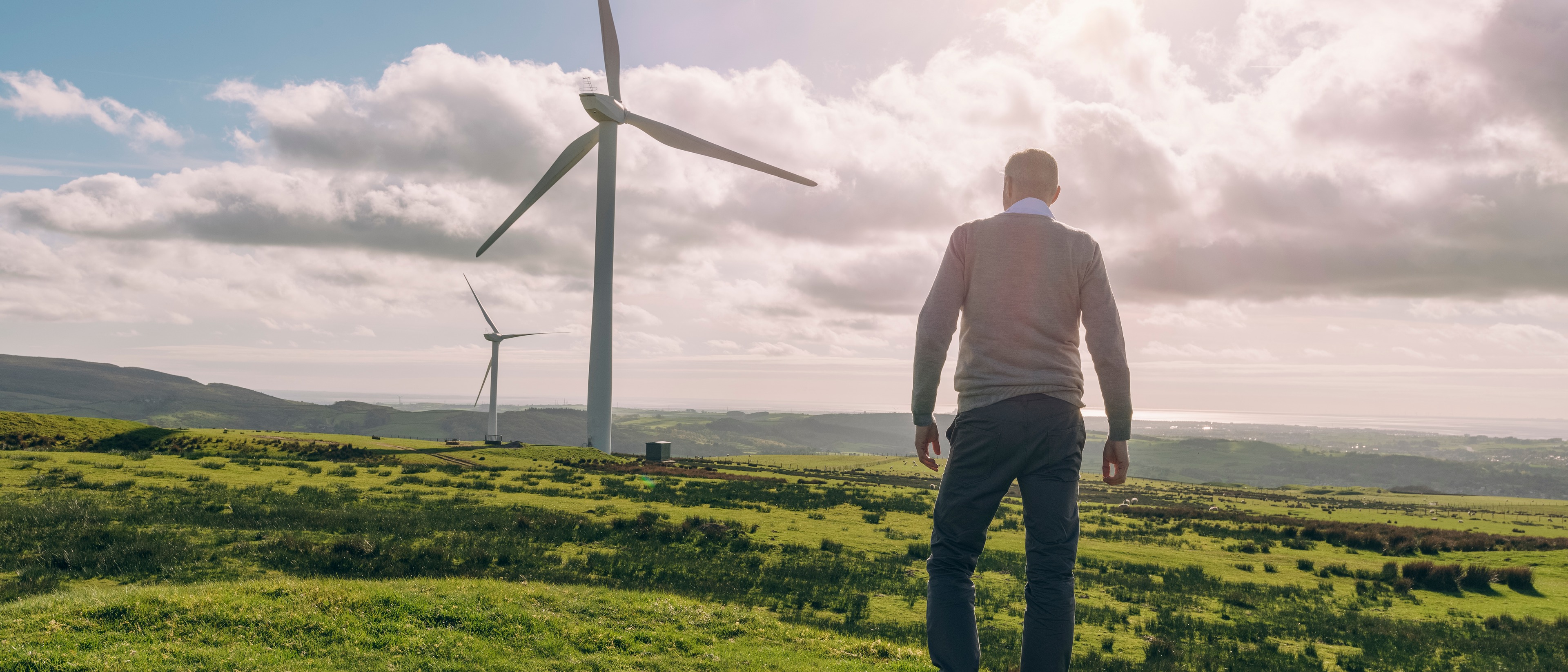 A man is standing in the landscape and is looking into the distance, at two wind turbines