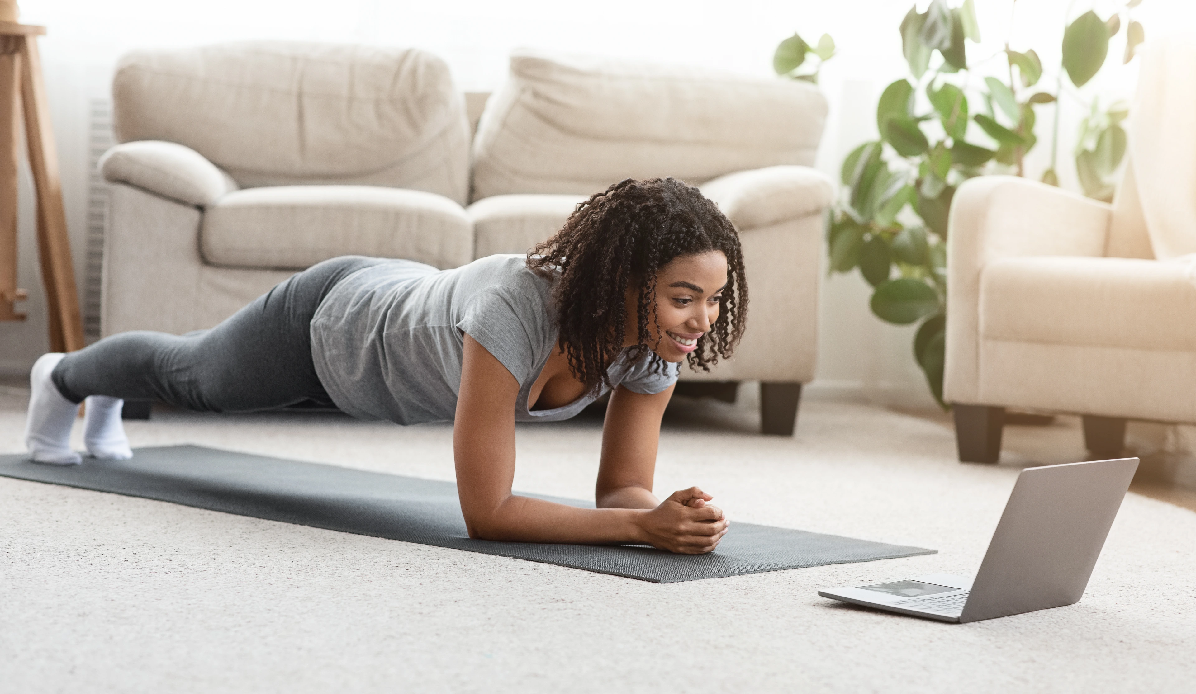 Training At Home. Fit black woman doing yoga plank while watching online tutorial on laptop, exercising in living room, free space, panorama