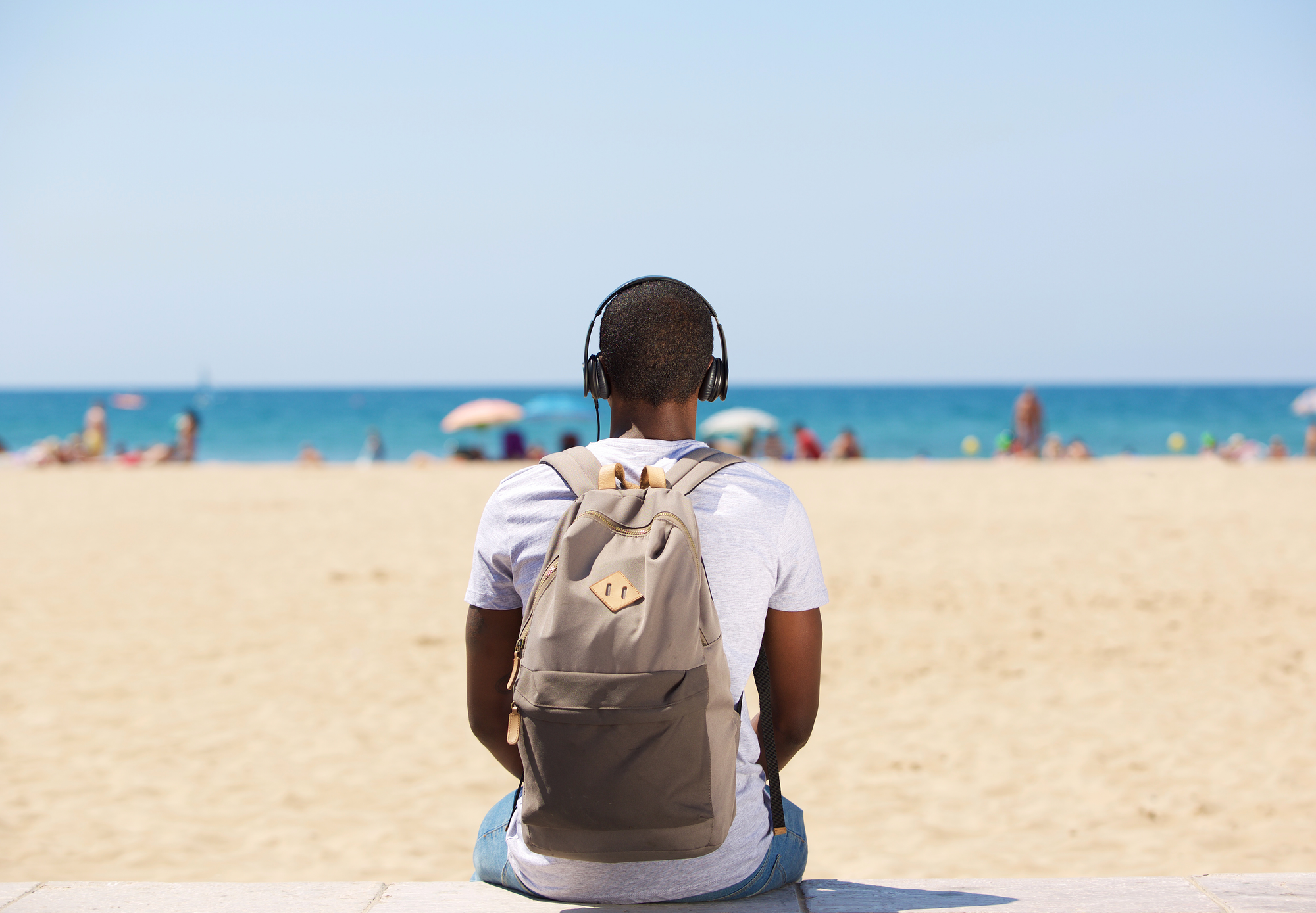 Person mit Rucksack und Kopfhörern sitzt am Strand und blickt aufs Meer, im Hintergrund sind Sonnenschirme und Badegäste zu sehen.