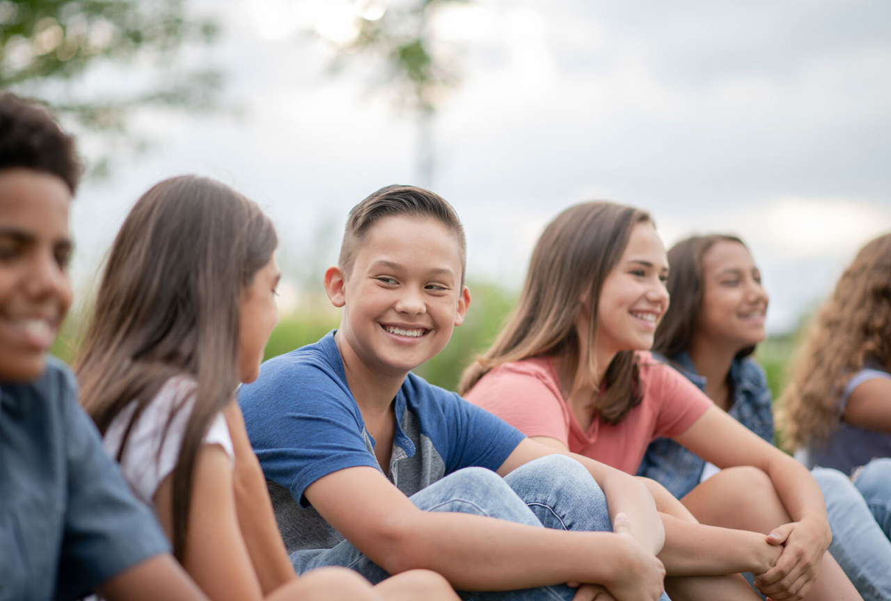 Eine Gruppe von Kindern bzw. Teenagern, die draußen auf dem Rasen sitzen, fröhliche Gesichter, verschiedene Geschlechter und Hautfarben