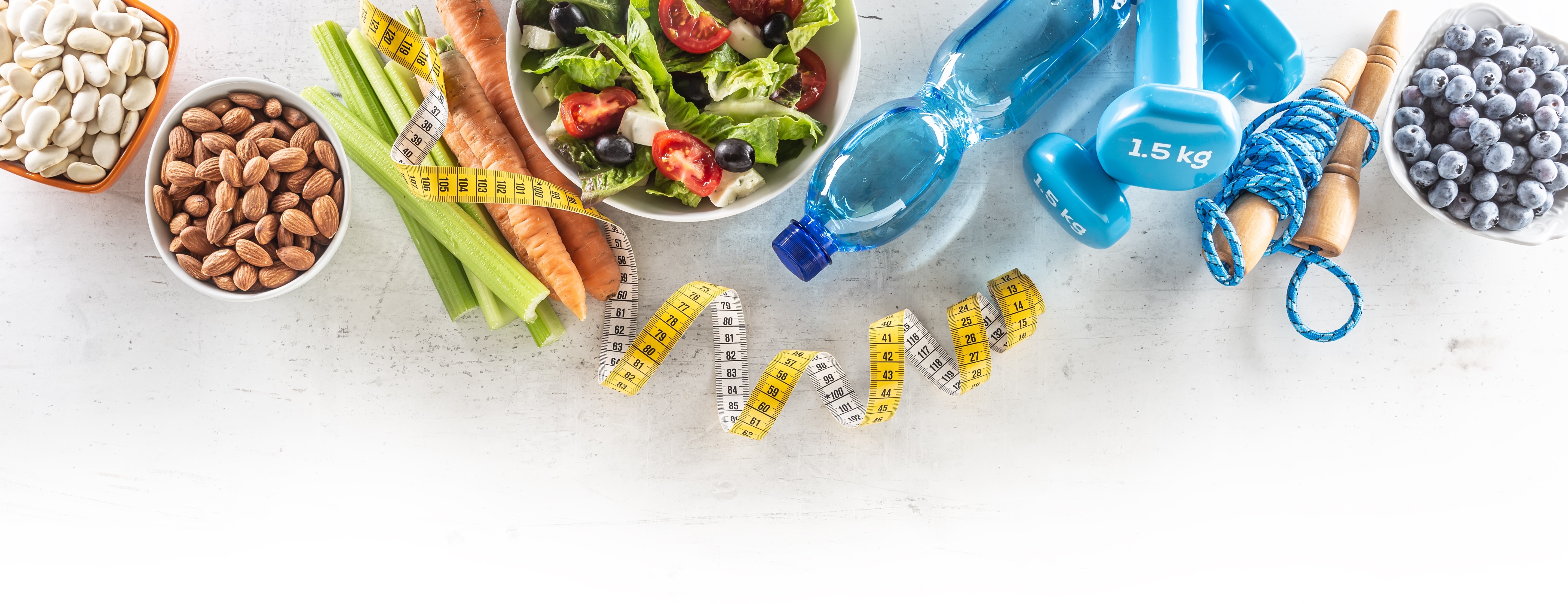 Healthy fresh salad with tomatoes surrounded with exercise equipment, carrtot celery and measuring tape - top of view.