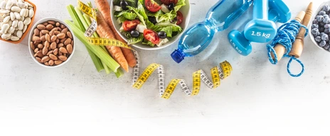 Healthy fresh salad with tomatoes surrounded with exercise equipment, carrtot celery and measuring tape - top of view.
