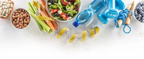 Healthy fresh salad with tomatoes surrounded with exercise equipment, carrtot celery and measuring tape - top of view.
