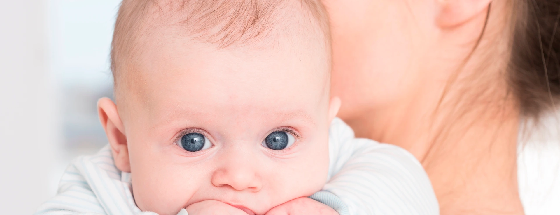 Little cute baby with big blue eyes sucking his little fist in mouth. Lying on mother's arm