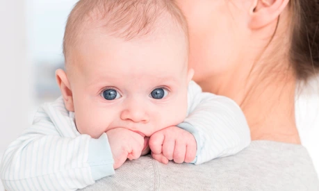 Little cute baby with big blue eyes sucking his little fist in mouth. Lying on mother's arm