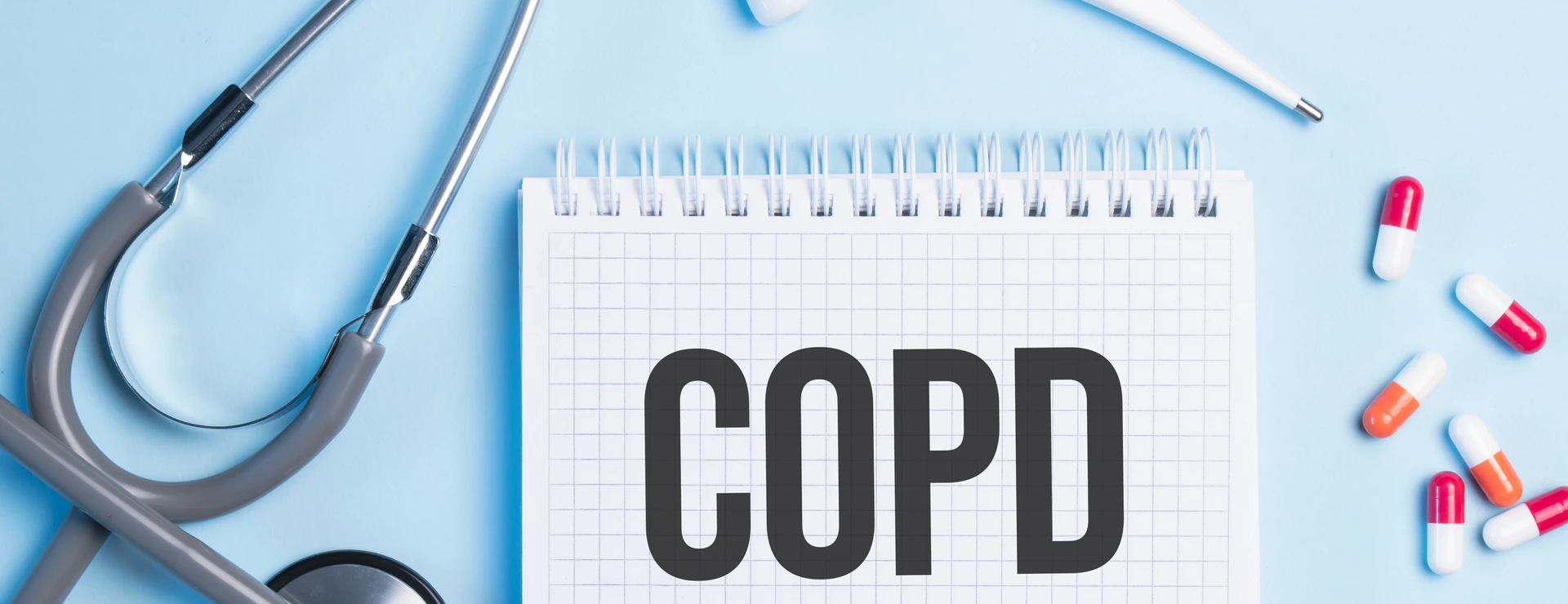 The word copd written on a white notepad on a blue background near a stethoscope, syringe, electronic thermometer and pills. Medical concept