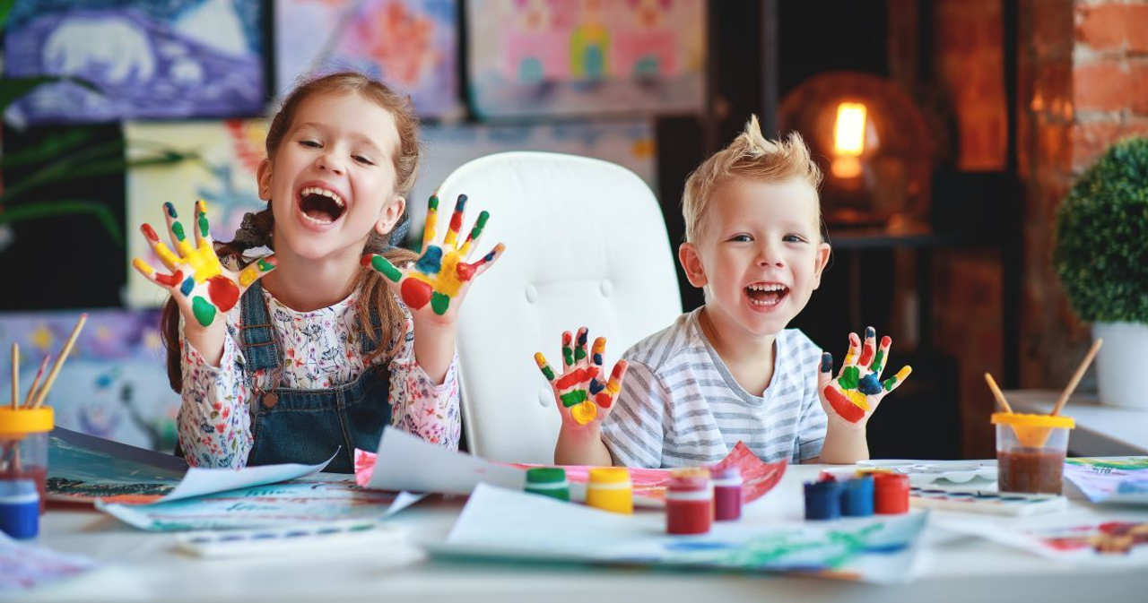 Duas crianças mostram as mãos pintadas enquanto fazem trabalhos de pintura numa mesa.