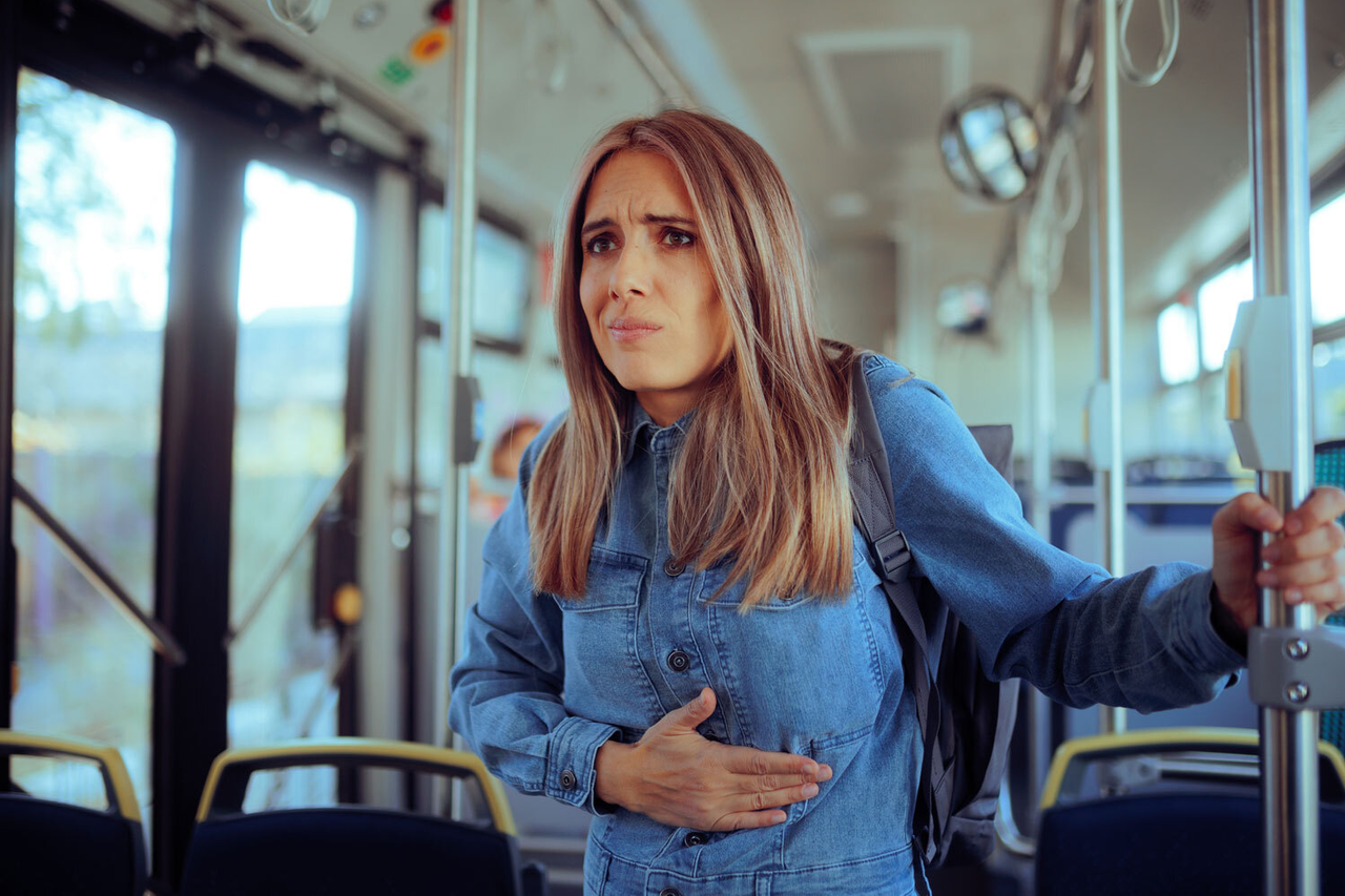 Foto einer jungen Frau in einem Bus, die das Gesicht verzieht, als ob ihr übel ist. Sie hält sich mit einer Hand den Bauch fest und sieht unglücklich aus.