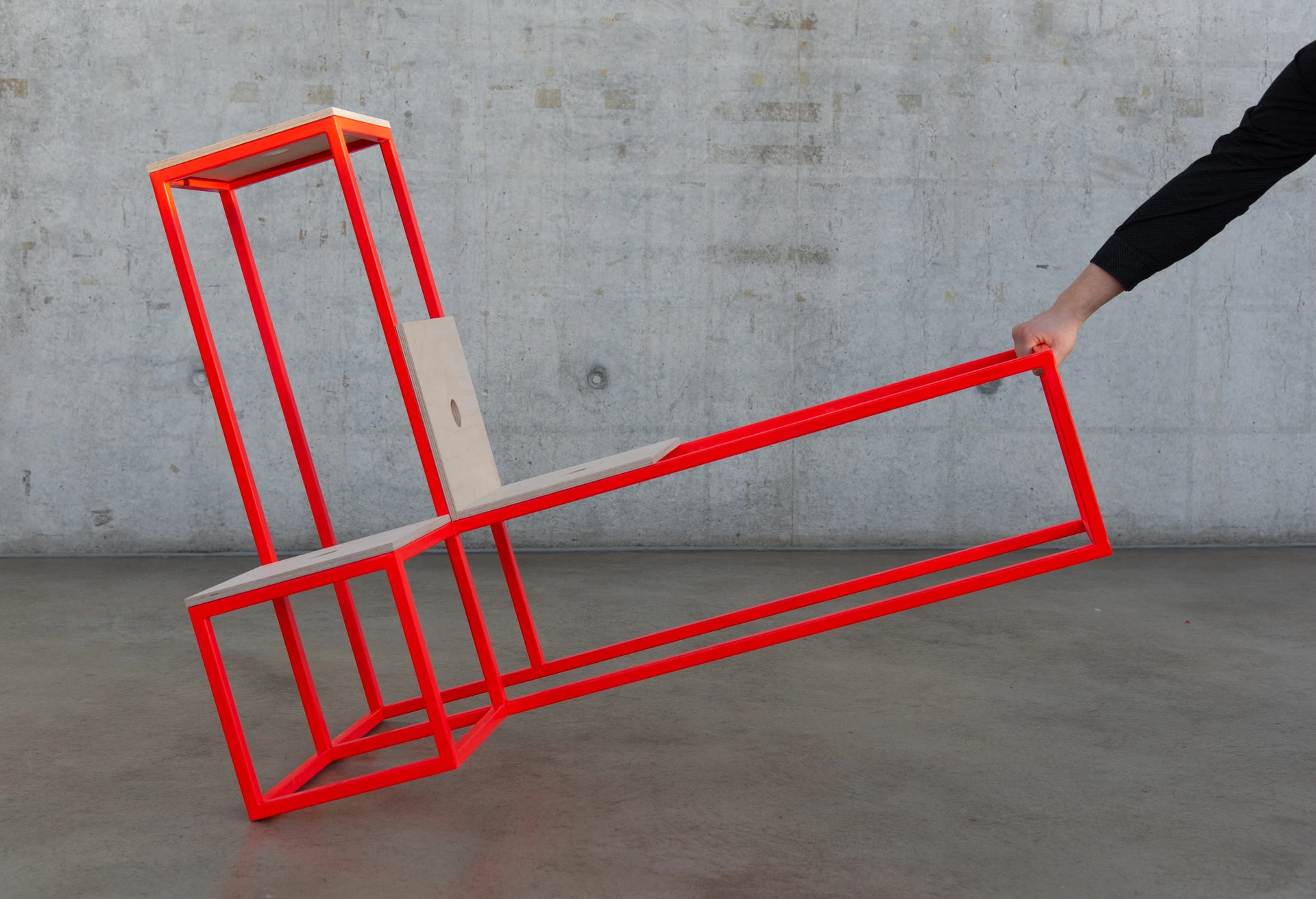 Designstuhl Salto von PALMBERG mit rotem Metallrahmen und Holzsitz, wird von einer Person in schräge Position gekippt, vor Betonwand.