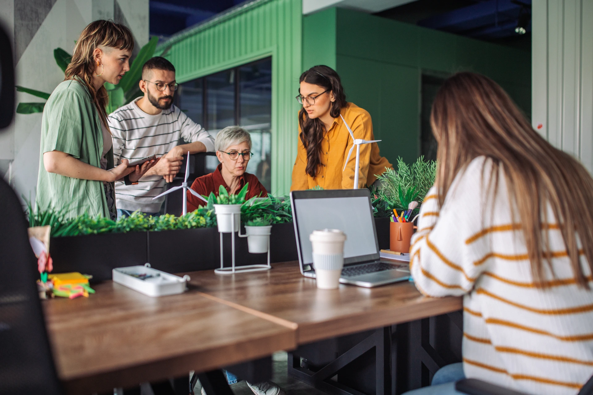 A group of people are engaged in teamwork, discussing around a laptop on a wooden table in a modern office space, emphasizing collaboration and creativity.