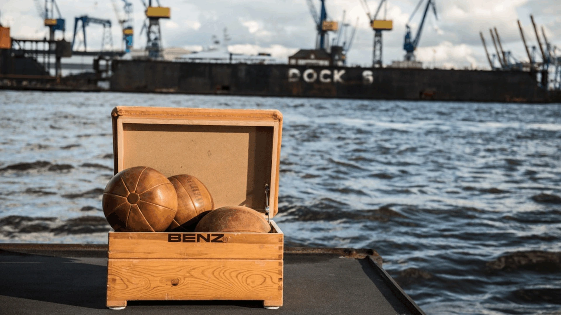 Holzkiste mit BENZ-Schrift und drei Lederbällen auf dem Kai, Frachtschiff im Hintergrund, Foto von PALMBERG