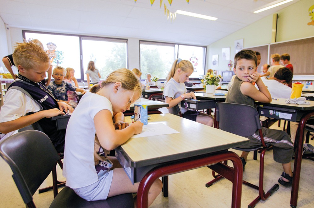 rundschulkinder sitzen in einem Klassenraum an ihren Tischen; einige schreiben oder malen, andere beschäftigen sich mit Materialien, während ein Junge seitlich den Blick durch den Raum wirft.