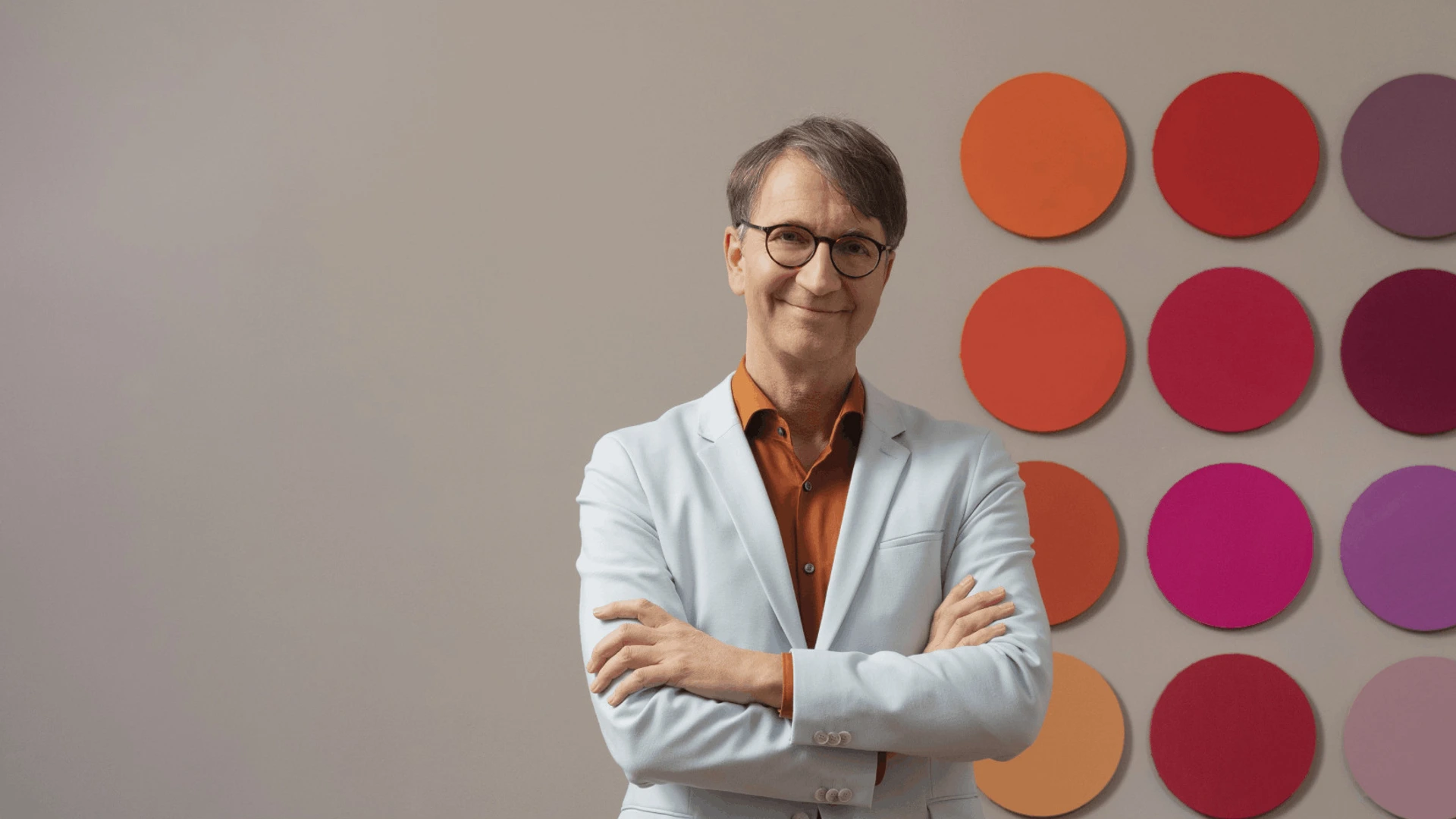 Lächelnder Mann mit Brille und verschränkten Armen vor bunter, kreisförmiger Wandinstallation von PALMBERG