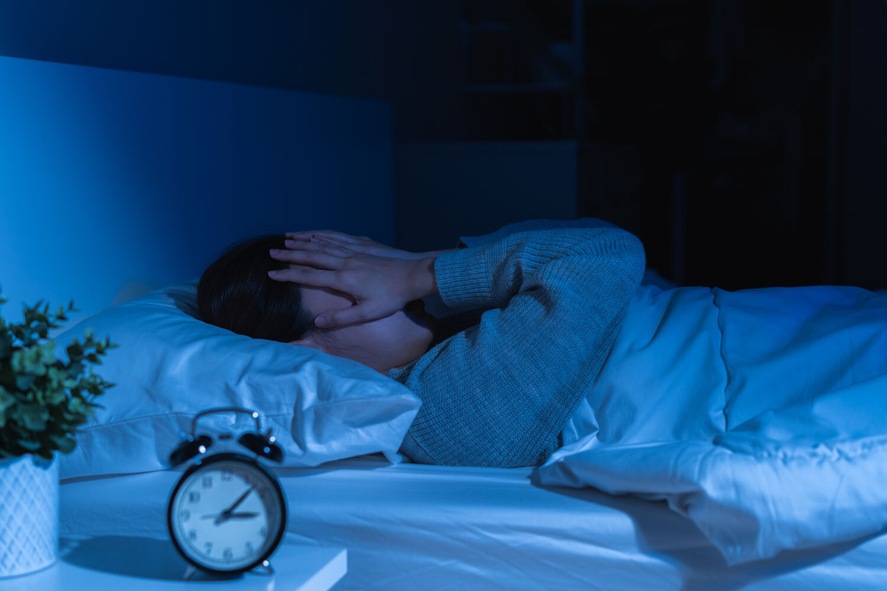 Eine Person liegt im Bett, im Vordergrund ein Wecker, der auf 10 Minuten nach 3 Uhr steht. Der Mensch hält sich die Hände vors Gesicht