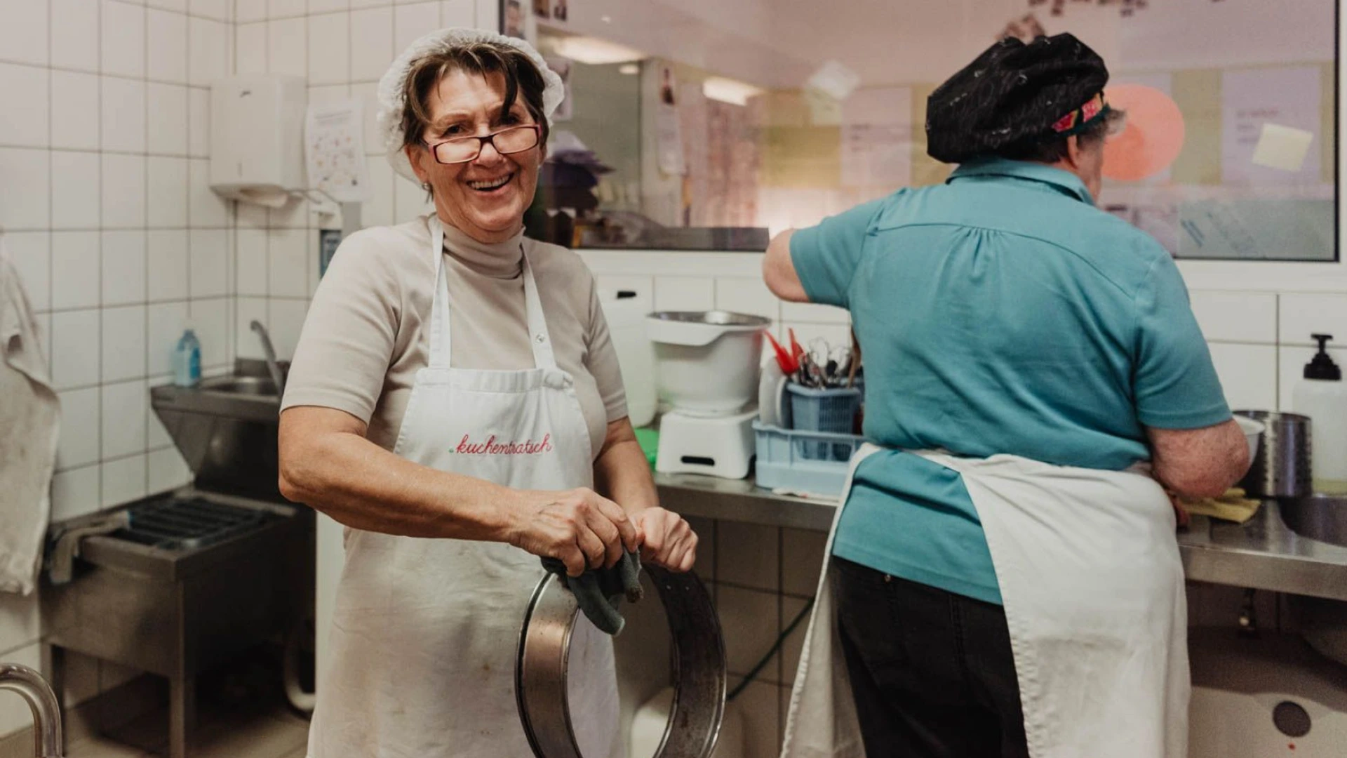 Zwei Frauen in Küchenkleidung, eine lächelt mit Sieb, Schürze mit dem Schriftzug kuchenback, Küche mit PALMBERG-Mobiliar