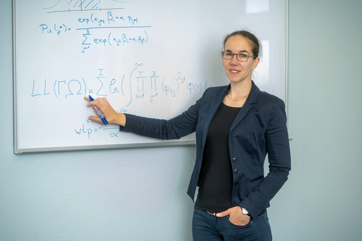 Eine Person steht an einer Tafel und zeigt auf mit der Hand geschriebene mathematische Formeln. 