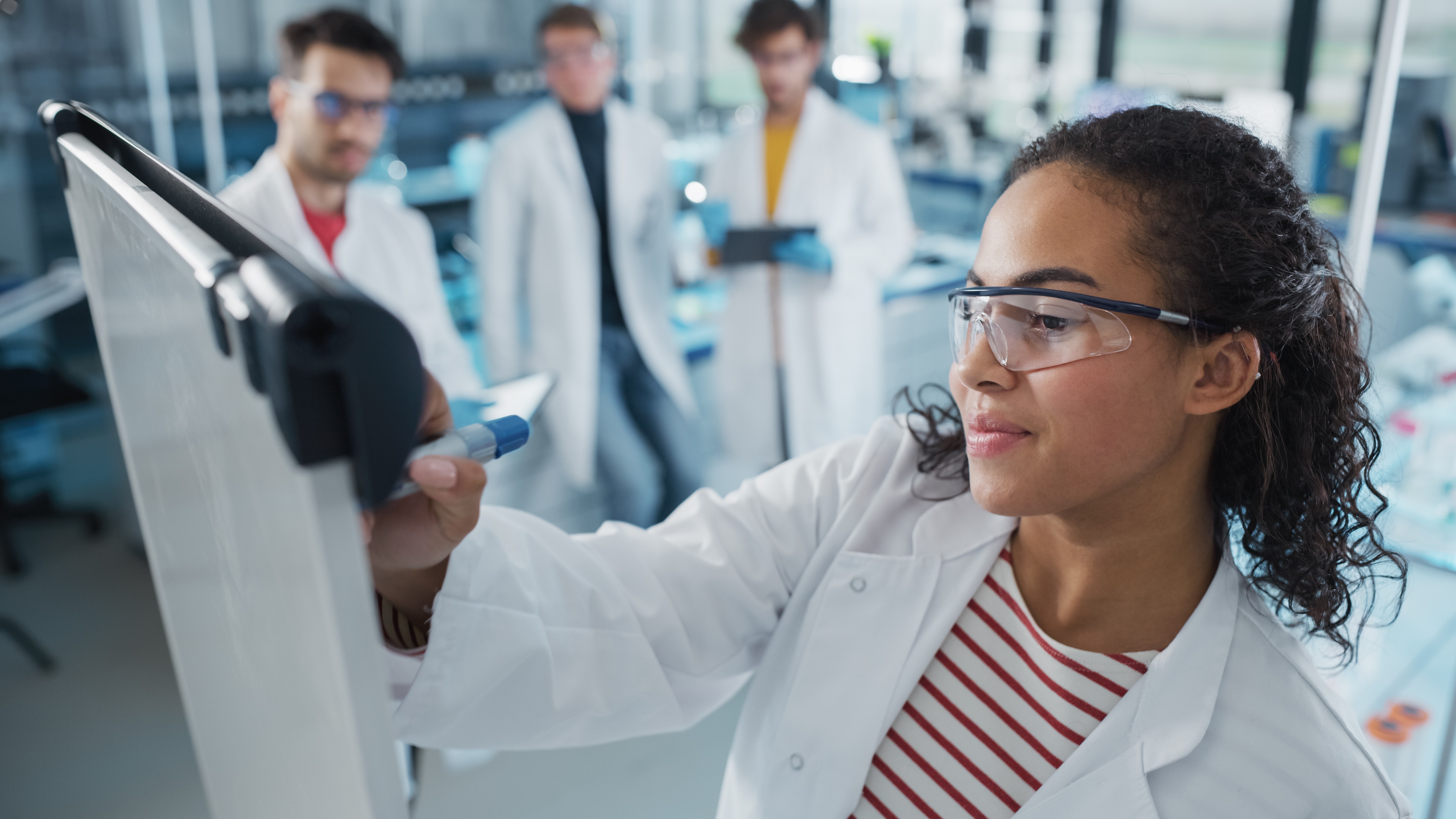 Medical Science Laboratory: Beautiful Black Female Scientist Writes Detailed Project Data Analysis on the Board, Her Diverse Team of Colleague Listens. Young Scientists Solving Problems.