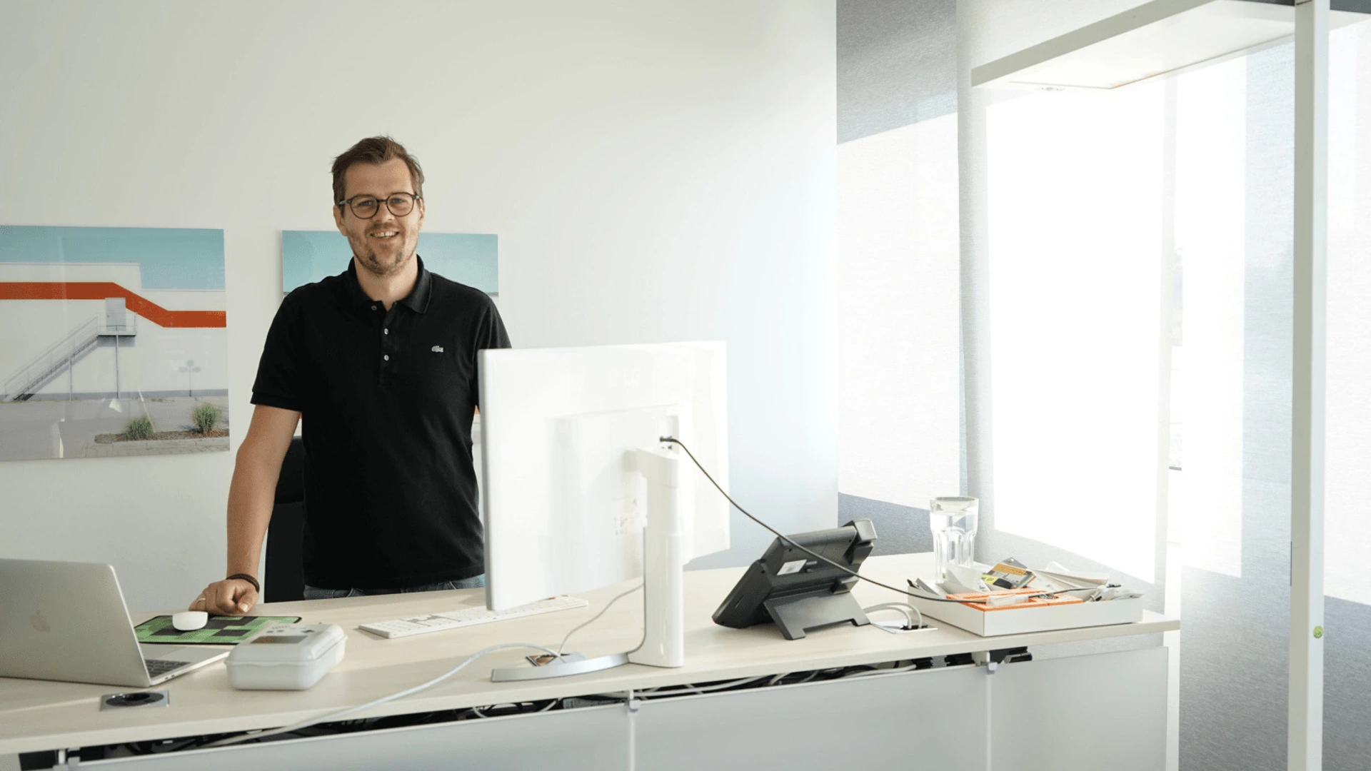 Mann steht lächelnd an einem Stehschreibtisch mit Monitor und Laptop, helle Büroumgebung, Büromöbel von PALMBERG
