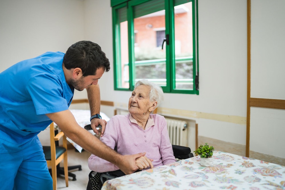 Eine männliche Pflegefachkraft wendet sich einer Seniorin zu, die an einem Tisch sitzt. Er schaut sie an und seine Hand hält ihre Hand. Sie schaut ihn an.