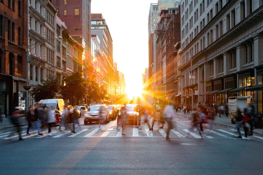 Crowds of busy people walking through the intersection of 5th Avenue and 23rd Street in Manhattan, New York City with bright sunset background