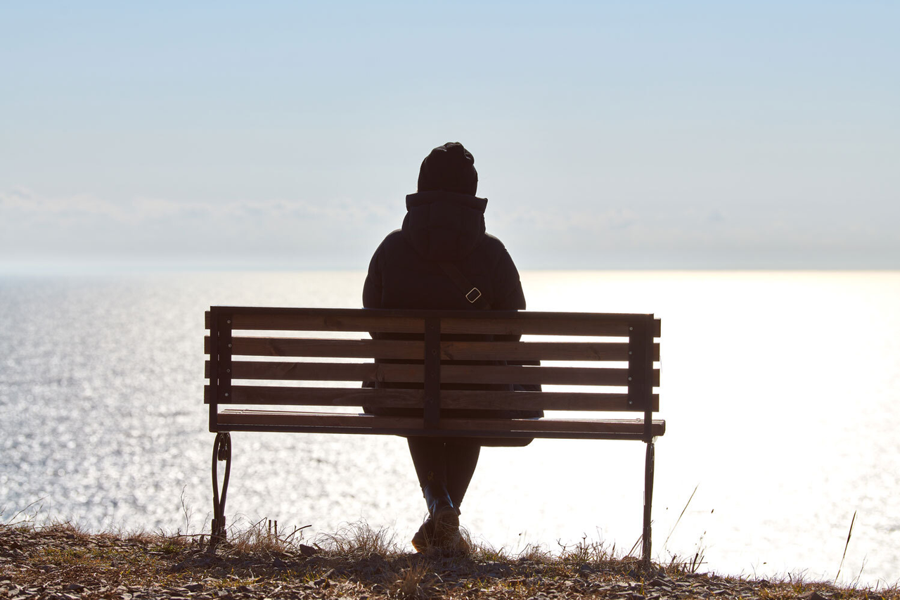Eine Frau, die allein auf einer Bank sitzt und aufs Meer schaut. Rückenansicht