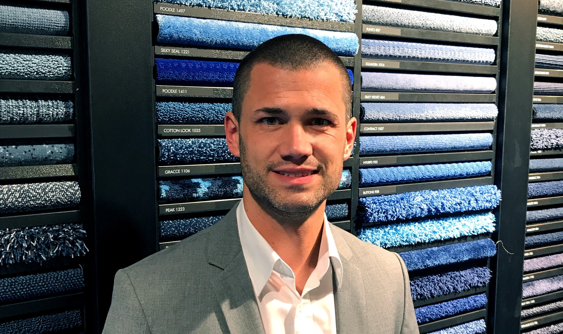 Lächelnder Mann im Anzug vor Wand mit blauen Teppichmustern und Beschriftung COTTON LOOK 1025, Produkte von PALMBERG