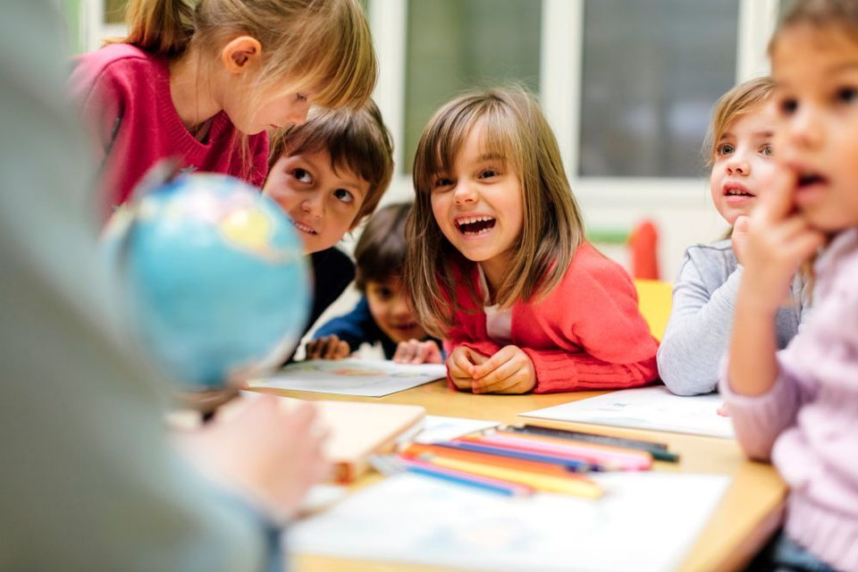 Gruppe von Grundschulkindern sitzt lachend und neugierig am Tisch im Klassenzimmer, während sie einer Person mit einem Globus zuhören.