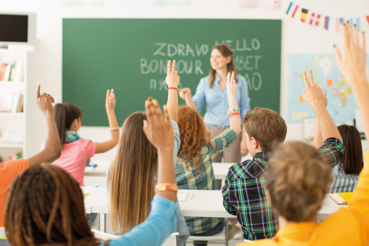 Schülerinnen und Schüler sitzen in einem Klassenzimmer, heben die Hände und melden sich, während eine Lehrerin vor der Tafel mit mehrsprachigen Begrüßungen steht.